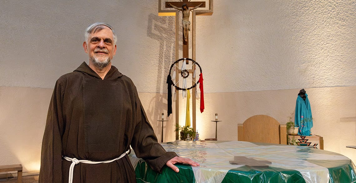 Photo of Br. Jozef Timmers in his brown Capuchin habit standing with his hand on the altar at St. Labre. A crucifix is behind him along with a Native American dreamcatcher.